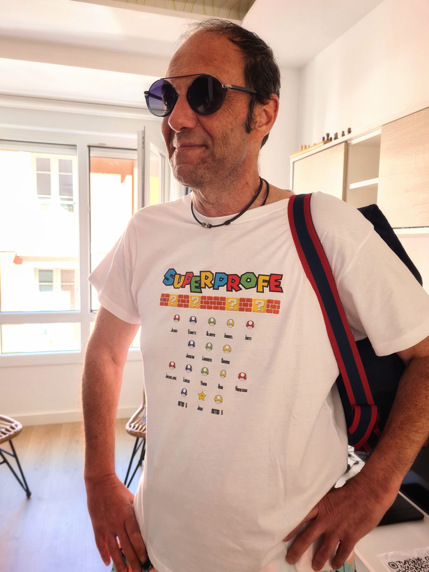 un hombre con gafas de sol y una camiseta blanca con texto colorido y pequeños iconos se encuentra de pie en una habitación luminosa, con una ventana y estanterías repletas de artículos como bragas al fondo. lleva un bolso azul marino con correas rojas sobre el hombro.